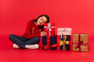 Noel hediyelerinin yanında kışın süveter ve kot pantolon giyen mutlu bir çocuk oturuyor.