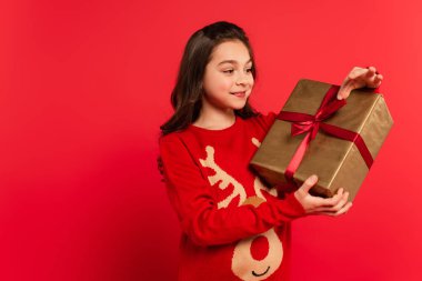 Mutlu çocuk örgü süveter içinde kırmızı üzerinde paketlenmiş Noel hediyesine bakıyor.