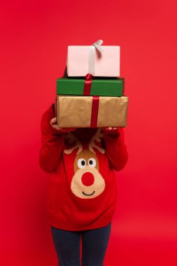 Kışın süveter giyen çocuk elinde kırmızı üzerine izole edilmiş bir sürü Noel hediyesi tutuyor.