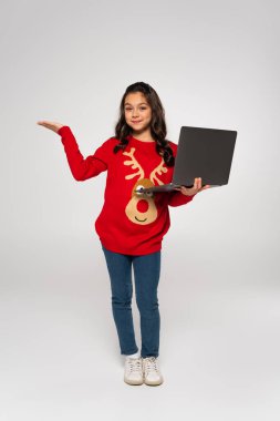 full length of joyful child in red sweater holding laptop and pointing with hand on grey clipart