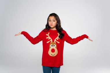 Preteen girl in festive sweater showing shrug gesture isolated on grey  clipart