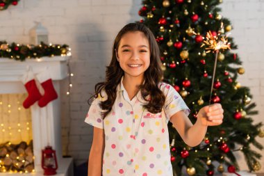 Pijamalı gülümseyen kız maytap tutuyor ve evde Noel kutlaması sırasında kameraya bakıyor. 