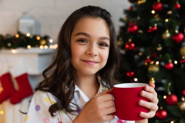 Pijamalı bir kız elinde fincanla evde bulanık Noel ağacının yakınındaki kameraya bakıyor. 