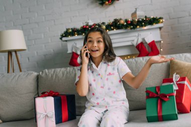 Heyecanlı çocuk akıllı telefonuyla konuşuyor Noel hediyeleri yanında evdeki koltukta 