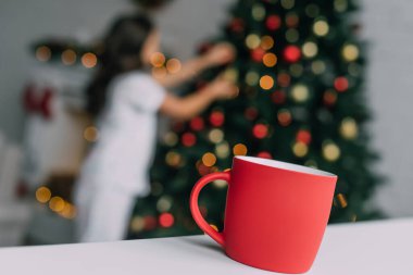 Red cup on table near blurred girl and Christmas tree at home  clipart