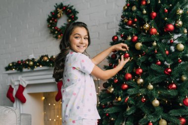 Evde Noel boyunca pijamalı, çam ağacını süsleyen gülümseyen kız. 