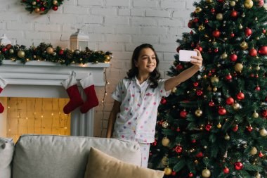 Pijamalı kız şöminenin yanındaki akıllı telefondan selfie çekiyor ve evde Noel ağacı var. 