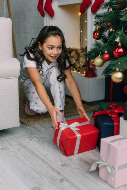 Pijamalı neşeli çocuk oturma odasındaki Noel ağacının altında hediye alıyor. 
