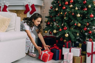 Pijamalı gülümseyen kız oturma odasındaki Noel ağacının altında hediye alıyor. 