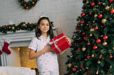 Evde Noel ağacının yanında elinde hediyeyle kameraya bakan pijamalı bir çocuk. 