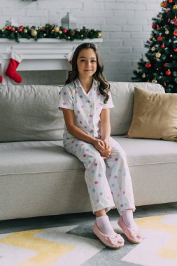Noktalı pijamalı gülümseyen kız bulanık şöminenin yanındaki koltukta oturuyor ve evde Noel ağacı var. 