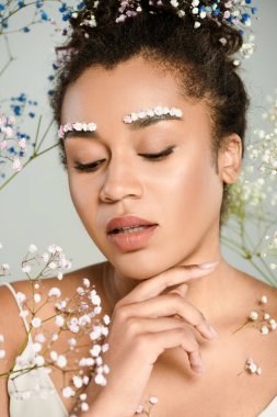 young african american woman with flowers in hair looking down near gypsophila isolated on grey clipart