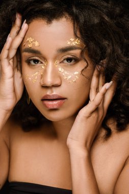 curly african american woman with golden paint on face looking at camera isolated on black clipart
