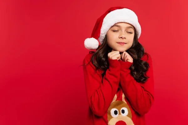 happy girl with closed eyes standing in santa hat and Christmas sweater isolated on red