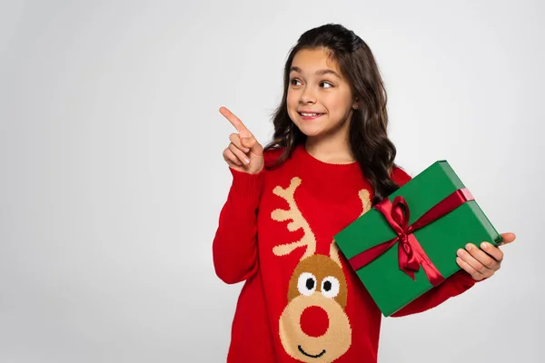 Uśmiechnięta Dziewczyna Świątecznym Swetrze Trzyma Prezent Wskazuje Palcem Szarym — Zdjęcie stockowe