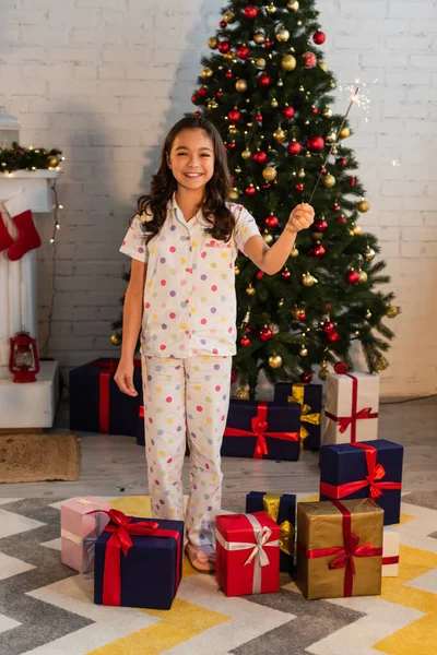 睡衣里的少女拿着火花 看着家里礼物和圣诞树旁边的相机 — 图库照片