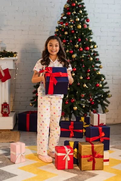 圣诞期间 坐在睡衣里快乐的孩子拿着礼品盒 看着相机 — 图库照片