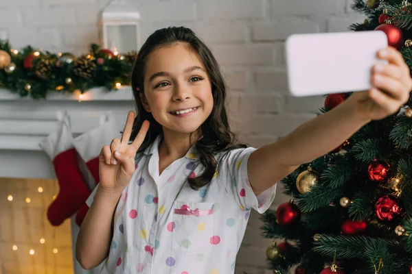 Smiling girl in pajama showing peace gesture while taking selfie on smartphone near Christmas tree