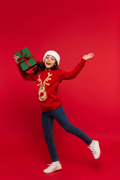 身穿圣诞毛衣 头戴圣诞礼帽 头戴红色背景礼品盒的快乐女孩 — 图库照片