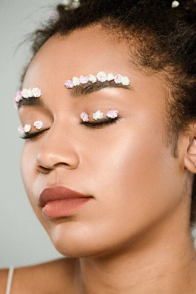 close up view of pretty african american woman with flowers on eyebrows and closed eyes isolated on grey