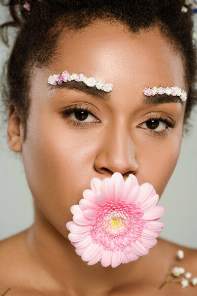 close up of african american woman with flowers on eyebrows and hair with gerbera in mouth isolated on grey