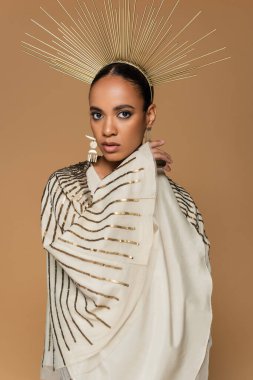 Parlak şallı ve altın kaplamalı Afro-Amerikan kadını bej renkli kameraya bakıyor. 