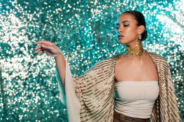 Parlak mavi arka planda altın rengi şalla poz veren, piercing takan Afro-Amerikan modeli. 