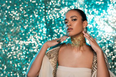 young african american woman in shawl and gold on neck looking at camera on sparkling blue background  clipart