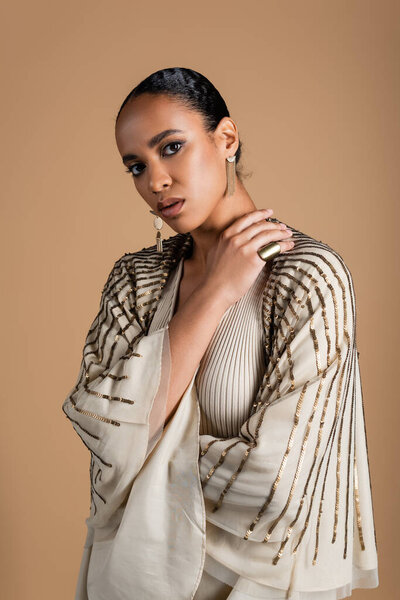 брюнетка африканская американка в золотых украшениях смотрит на камеру, изолированную от бежевого