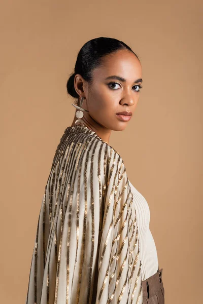 young african american model in golden jewelry looking at camera isolated on beige