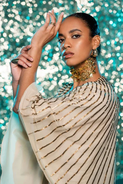 pierced african american model with jewelry and gold foil on neck posing while looking at camera on shiny blue background