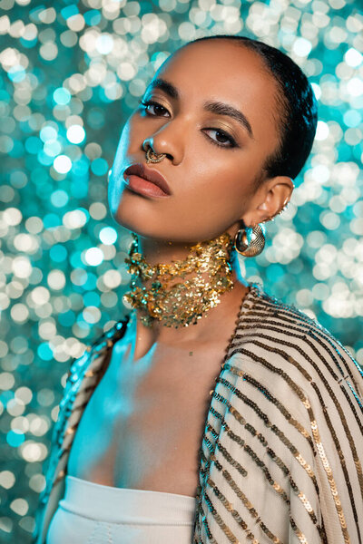 african american model with piercing and gold on neck posing while looking at camera on shiny blue background 