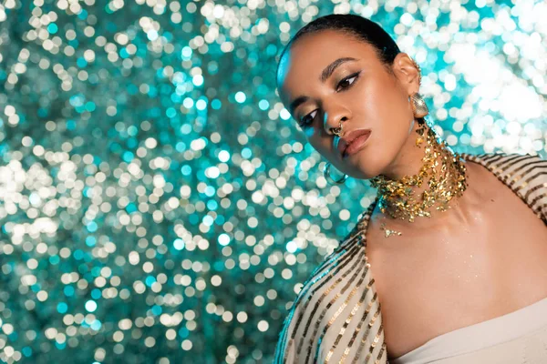 african american model with piercing and gold on neck posing while looking away on shiny blue background