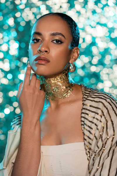 pierced african american woman in earrings and gold on neck posing on sparkling blue background