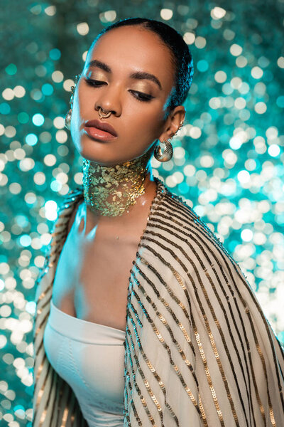 pierced african american woman with jewelry and gold on neck posing on sparkling blue background 