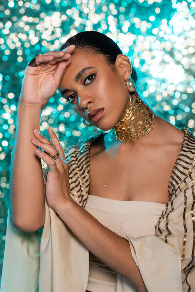 young african american woman in shawl and gold on neck looking at camera while posing on sparkling blue background 