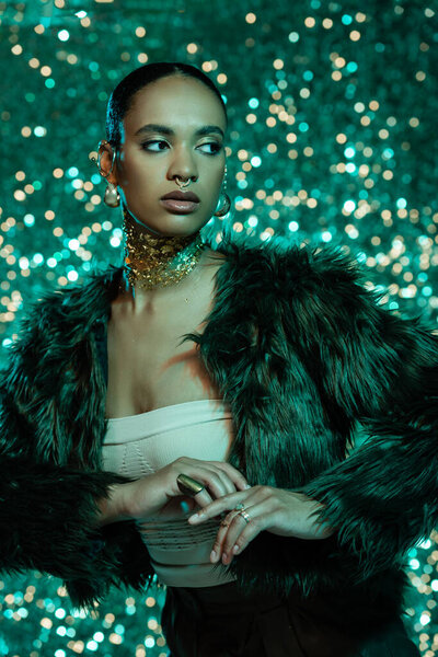 brunette african american woman in faux fur jacket and gold on neck posing on sparkling turquoise background 