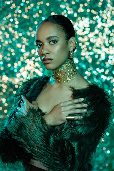 stock image young african american woman in faux fur jacket and gold on neck posing with crossed arms on sparkling turquoise background 