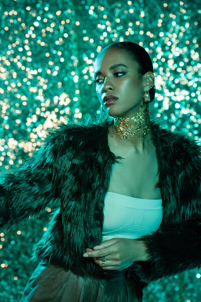 stylish african american model in faux fur jacket and gold on neck posing on shiny blue background 