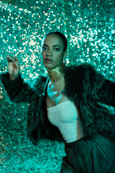 trendy african american woman in faux fur jacket and gold on neck posing on shiny blue background 