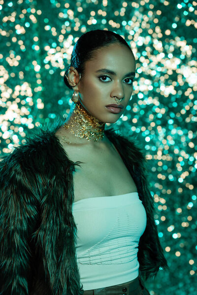young african american woman in trendy faux fur jacket and gold on neck looking at camera on shiny blue background 