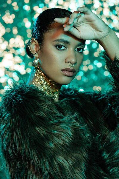 Stylish african american woman in faux fur jacket looking at camera on sparkling background 