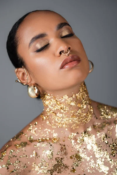 African american model with golden accessories and foil on body closing eyes isolated on grey