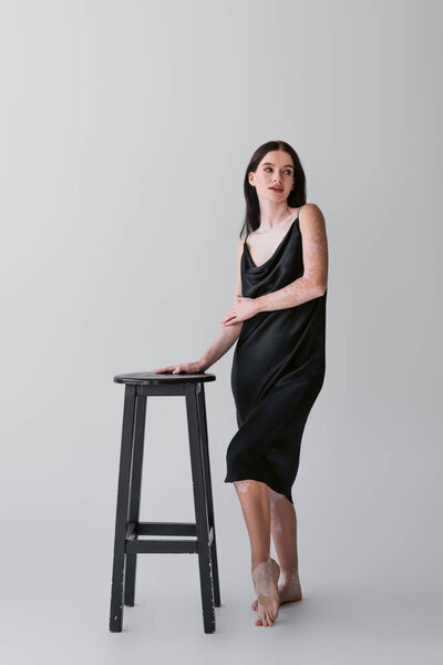 Full length of young woman with vitiligo looking away near chair on grey background 