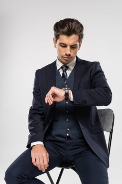 Resmi olarak genç işadamı, gri üzerine izole edilmiş kol saatine bakarken sandalyede oturuyor.