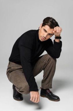Siyah balıkçı yaka kazaklı esmer adam gri üzerinde poz verirken oturuyor.