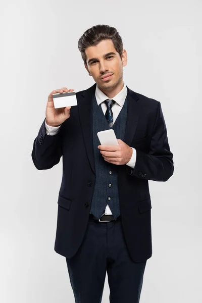 スーツ姿の若いビジネスマンがグレーで隔離されたスマートフォンやクレジットカードを持ち — ストック写真