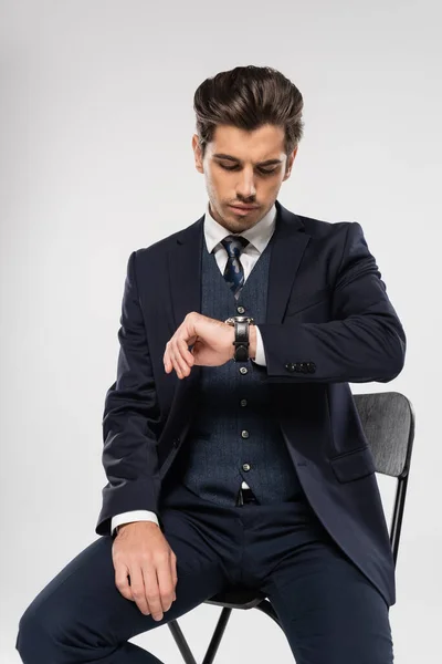 グレーで隔絶された腕時計を見ながら椅子に座る若い実業家 — ストック写真