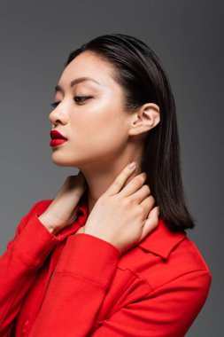 portait of asian woman in red jacket posing with hands on neck isolated on grey clipart