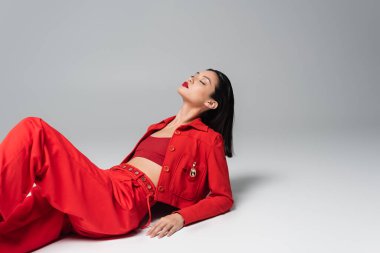 Şehvetli Asyalı kadın. Gözleri kapalı. Gri arka planda kırmızı şık kıyafetler giyiyor.